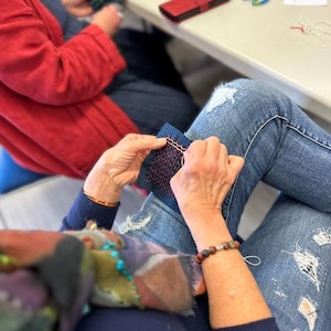 May include: A person is hand-stitching a small, rectangular piece of dark blue fabric with a grid of white dots. The fabric features a row of pink wavy embroidery stitches at the top. The person is wearing blue distressed denim jeans and a dark sweater.