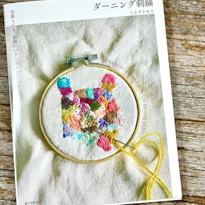 Può includere: Un telaio da ricamo con un disegno di gatto cucito a colori. Il ricamo è su tessuto bianco, con filo e ago gialli. Il telaio è posto su un libro con testo giapponese.