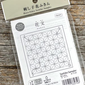 Pode incluir: Um kit Hana-Fukin Sashiko Sampler em embalagem plástica transparente. O tecido branco apresenta um padrão floral com linhas pontilhadas para costura. O pacote inclui instruções em inglês e o tamanho final é de aproximadamente 34 x 34 cm.