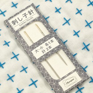 May include: A package of two sashiko needles, one large and one small, from Olympus. The package is gray with a white border and features a repeating pattern of circles. The text on the package reads "刺し子針 Sashiko Needle" and "大・小各1本 計2本".