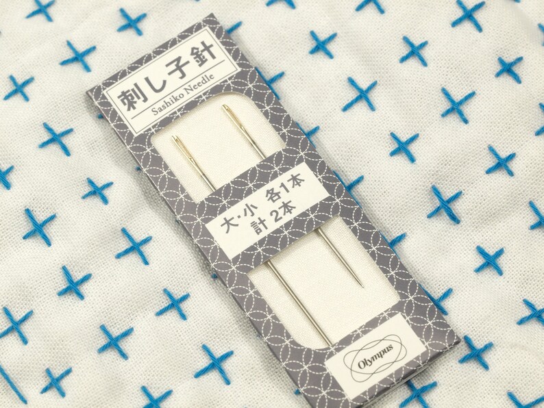 May include: A package of two sashiko needles, one large and one small, from Olympus. The package is gray with a white border and features a repeating pattern of circles. The text on the package reads "刺し子針 Sashiko Needle" and "大・小各1本 計2本".