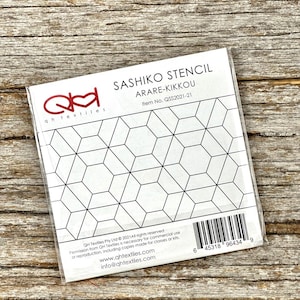 May include: A clear plastic stencil with a geometric pattern of interconnected hexagons and triangles. The stencil is labeled "SASHIKO STENCIL ARARE-KIKKOU" with the item number QS$2021-21. The stencil is on a wooden surface.