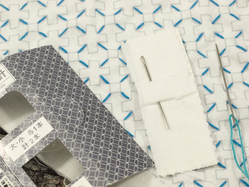 May include: A close-up of a white fabric with blue stitching in a cross pattern. Two silver needles are on a white paper with the text "大・小 各1本 計2本".