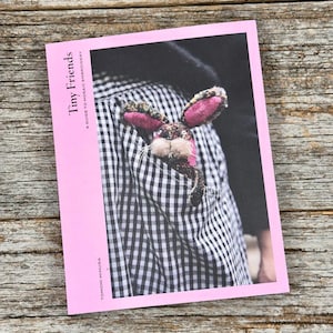 May include: A pink book titled "Tiny Friends" with the subtitle "A Guide to Mosaic Embroidery" by Tomomi Mimura. The book's cover features a photo of a stuffed bunny peeking out of a pocket on a black and white checkered shirt.
