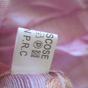 May include: A close-up of a white clothing label with black text and care symbols. The label is attached to a pink and white striped fabric. The label reads "SCOSE N.P.R.C."