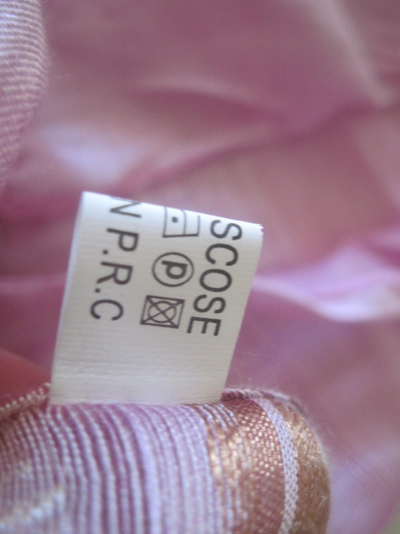May include: A close-up of a white clothing label with black text and care symbols. The label is attached to a pink and white striped fabric. The label reads "SCOSE N.P.R.C."
