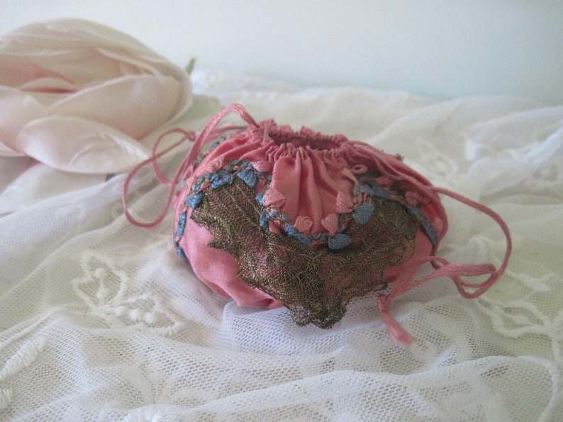 Op de afbeelding: Een kleine, roze trekkoordzak met blauwe en gouden accenten. De zak is gemaakt van een zachte stof en heeft een delicaat kanten ontwerp. Het trekkoord heeft een bijpassende roze kleur. De zak wordt waarschijnlijk gebruikt voor het dragen van kleine items.