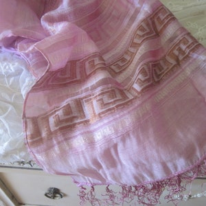 May include: A pale pink scarf with a geometric design in brown and gold. The scarf has a delicate fringe along the edge. The fabric appears to be lightweight and flowing, suitable for a shawl or wrap.