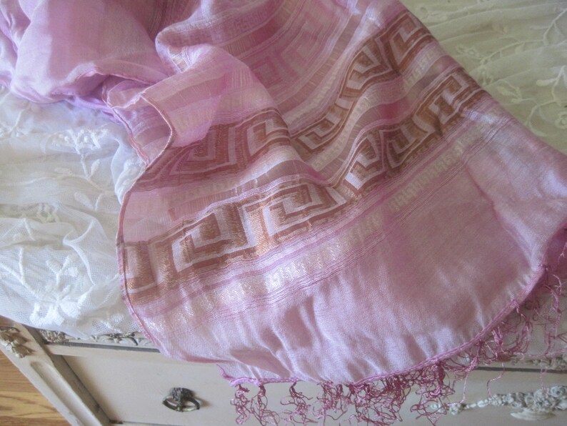 May include: A pale pink scarf with a geometric design in brown and gold. The scarf has a delicate fringe along the edge. The fabric appears to be lightweight and flowing, suitable for a shawl or wrap.