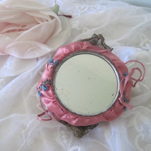 Op de afbeelding: Een vintage ronde handspiegel met een roze satijnen frame en een decoratieve metalen filigraan achterkant. De spiegel is versierd met kleine blauwe bloemaccenten en roze strikjes. Op de achtergrond ligt een grote, lichtroze roos.