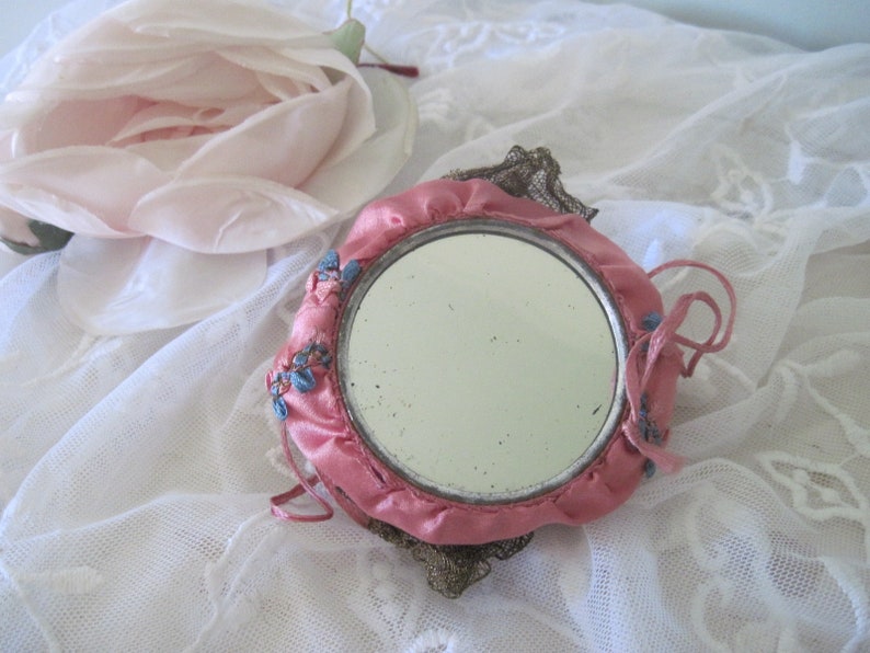 Op de afbeelding: Een vintage ronde handspiegel met een roze satijnen frame en een decoratieve metalen filigraan achterkant. De spiegel is versierd met kleine blauwe bloemaccenten en roze strikjes. Op de achtergrond ligt een grote, lichtroze roos.
