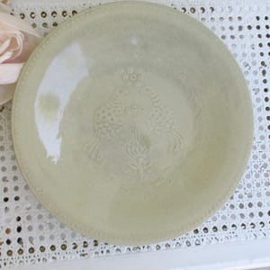 Pode incluir: Um prato de cerâmica redondo em verde sálvia pálido com um design central em relevo de dois pássaros voltados um para o outro acima de um motivo floral. A borda do prato é decorada com uma textura de contas sutil e acabamento brilhante.