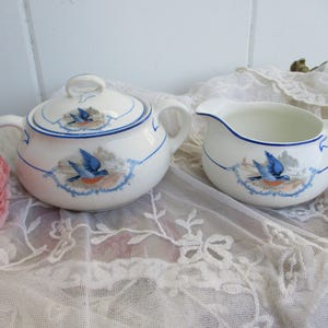 May include: A white ceramic sugar bowl with a lid and a creamer. Both pieces are decorated with a blue bird design and blue trim. The sugar bowl has two handles and a loop handle on the lid. The creamer has a handle and a spout.
