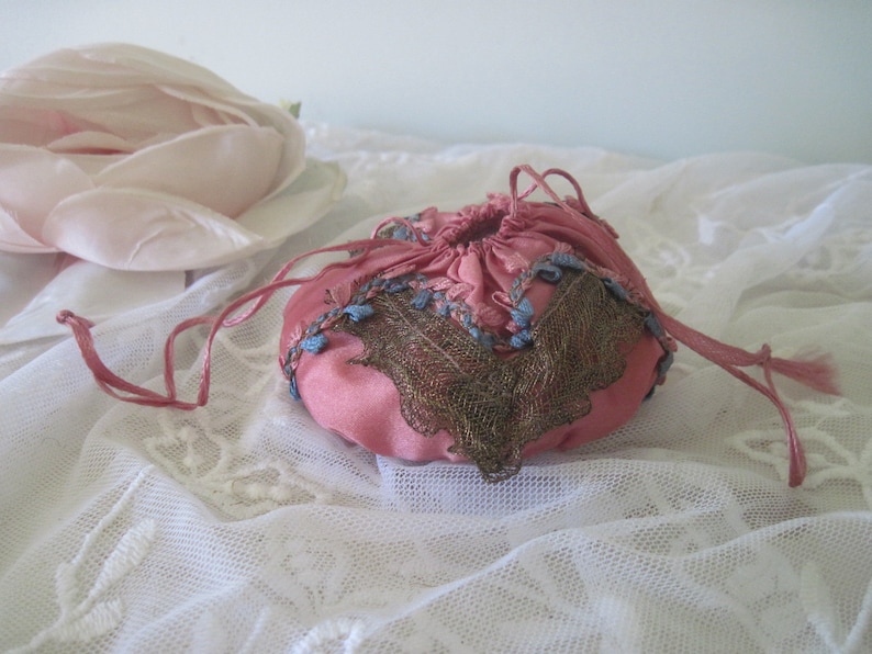 Op de afbeelding: Een kleine, roze satijnen zak met trekkoord en gedetailleerde borduursels. De zak heeft blauwe en gouden borduursels en een bijpassend roze trekkoord. Op de achtergrond een grote, lichtroze stoffen roos.
