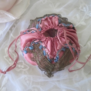 Op de afbeelding: Een vintage roze satijnen trekkoordtasje met blauwe en bruine decoratieve afwerking. Het tasje heeft een geplooid ontwerp en delicate blauwe bloemenborduurwerk. Roze linten zijn bevestigd om te sluiten en een kleine kwast hangt aan het trekkoord.