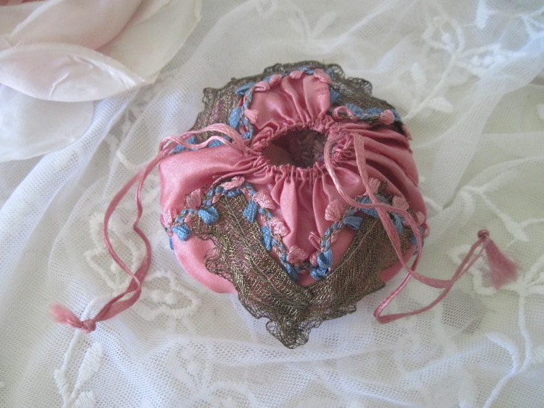 Op de afbeelding: Een vintage roze satijnen trekkoordtasje met blauwe en bruine decoratieve afwerking. Het tasje heeft een geplooid ontwerp en delicate blauwe bloemenborduurwerk. Roze linten zijn bevestigd om te sluiten en een kleine kwast hangt aan het trekkoord.