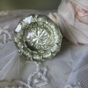 May include: A clear, faceted glass doorknob with a starburst design in the center. The knob has a vintage aesthetic and is likely from an antique door. The doorknob is a light green color and is sitting on a white lace surface.
