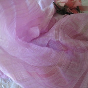 May include: A delicate, pale purple fabric with a subtle striped design. The fabric is draped, creating soft folds and shadows. A few pink roses are visible in the background, adding a touch of colour and texture. The fabric appears to be lightweight and fine.