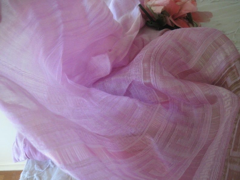 May include: A delicate, pale purple fabric with a subtle striped design. The fabric is draped, creating soft folds and shadows. A few pink roses are visible in the background, adding a touch of colour and texture. The fabric appears to be lightweight and fine.