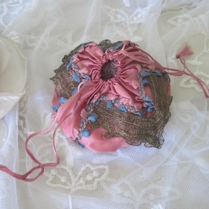 Op de afbeelding: Een kleine, ronde, roze trekkoordtas met blauw borduurwerk en een bronzen kanten rand. De tas is gemaakt van een zacht, mogelijk fluweelachtig materiaal en is vastgebonden met een roze koord. De achtergrond is een witte kanten stof.