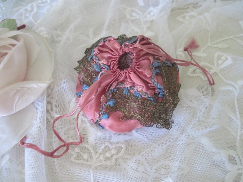 Op de afbeelding: Een kleine, ronde, roze trekkoordtas met blauw borduurwerk en een bronzen kanten rand. De tas is gemaakt van een zacht, mogelijk fluweelachtig materiaal en is vastgebonden met een roze koord. De achtergrond is een witte kanten stof.