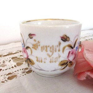 May include: A white porcelain cup with gold trim and floral accents. The cup features the words "Forget me not" in gold script. Pink and blue flowers are painted around the cup's exterior. A pink flower is in the background.