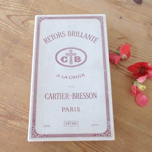 May include: A rectangular white box with burgundy text and a decorative border. The text reads "RETORS BRILLANTE A LA CROIX CARTIER-BRESSON PARIS." A sprig of red and pink flowers rests on a wooden surface.