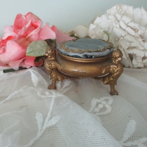 Puede incluir: Pequeña caja de baratijas dorada antigua con una parte superior de cojín de terciopelo azul y detalles ornamentados. La caja está sostenida por tres pies decorativos. Flores rosas y blancas en el fondo añaden un toque de elegancia.