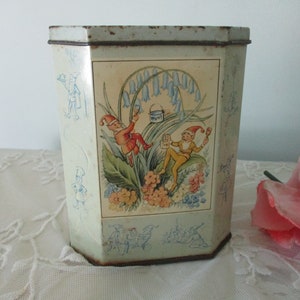 Peut inclure: Boîte en métal vintage de forme octogonale. L'illustration centrale représente deux gnomes dans un jardin avec des fleurs de jacinthes. Les côtés présentent des figures bleues esquissées. La boîte a un fond vert clair.