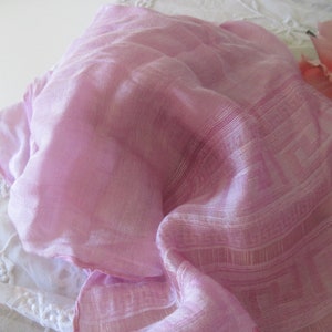 May include: A pale pink scarf with a delicate geometric design. The scarf is draped, revealing its fine texture and semi-transparent appearance. A pink rose is in the background, adding a touch of elegance.