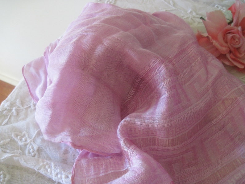 May include: A pale pink scarf with a delicate geometric design. The scarf is draped, revealing its fine texture and semi-transparent appearance. A pink rose is in the background, adding a touch of elegance.