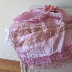 May include: A light pink scarf with a geometric pattern in a darker pink and gold. The scarf has a fringed edge and is draped over a white surface. A small bouquet of pink roses rests on top.