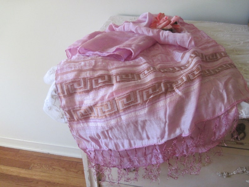 May include: A light pink scarf with a geometric pattern in a darker pink and gold. The scarf has a fringed edge and is draped over a white surface. A small bouquet of pink roses rests on top.