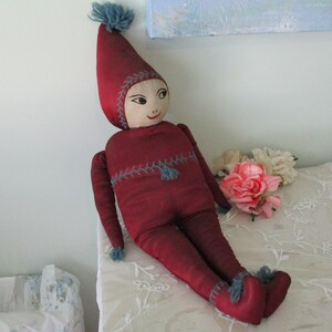Könnte beinhalten: Handgefertigte Puppe mit rotem Outfit und spitzem Hut. Die Puppe hat ein bemaltes Gesicht mit einem lächelnden Ausdruck und blauen Akzenten. Die Puppe ist etwa 30 cm groß und sitzt auf einer weißen Oberfläche.