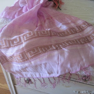 May include: A light pink scarf with a geometric border design in shades of brown. The scarf has a fringed edge and is draped over a white surface. The scarf is made of a lightweight, sheer fabric.