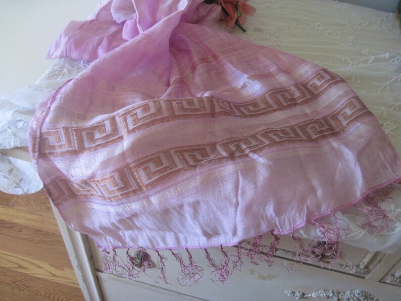 May include: A light pink scarf with a geometric border design in shades of brown. The scarf has a fringed edge and is draped over a white surface. The scarf is made of a lightweight, sheer fabric.