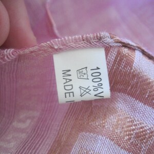 May include: Close-up of a pink and tan scarf with a label. The label reads "MADE IN" and "100% V" with a washing symbol. The scarf has a striped pattern and is made of a lightweight fabric.
