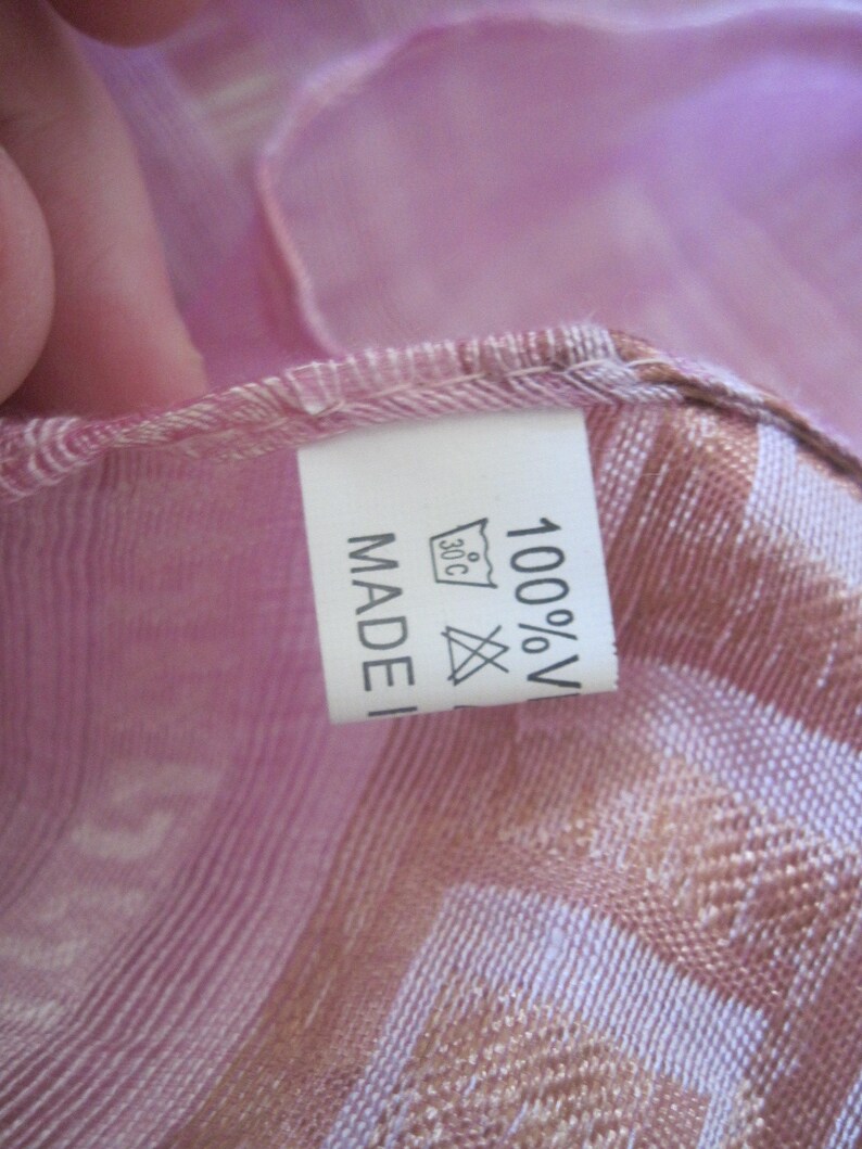 May include: Close-up of a pink and tan scarf with a label. The label reads "MADE IN" and "100% V" with a washing symbol. The scarf has a striped pattern and is made of a lightweight fabric.