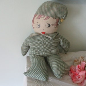 May include: A vintage cloth doll with a painted face, wearing a green and white polka dot outfit and matching cap. The doll has rosy cheeks, red lips, and a yellow flower accent on the cap. The doll is sitting on a white surface.