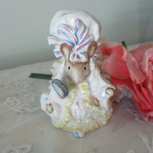 以下が含まれることがあります： 青とピンクのストライプの白い帽子と白いドレスを着たネズミの陶器人形。ネズミは小さな丸いものを持っています。人形はパステルカラーで手描きされています。