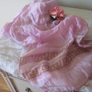 May include: A light pink scarf with a geometric border in shades of tan and pink. A pink rose rests on the scarf, which is draped over a white, embroidered surface. The scarf's fabric appears soft and lightweight.