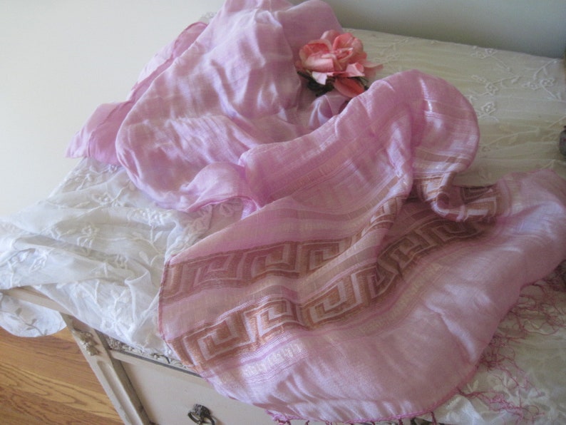 May include: A light pink scarf with a geometric border in shades of tan and pink. A pink rose rests on the scarf, which is draped over a white, embroidered surface. The scarf's fabric appears soft and lightweight.