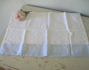 Op de afbeelding: Witte kussensloop met een blauwe gingham en oogjesrand. Een roze bloem ligt in de linkerbovenhoek van de kussensloop. De kussensloop is rechthoekig en lijkt van katoen te zijn.