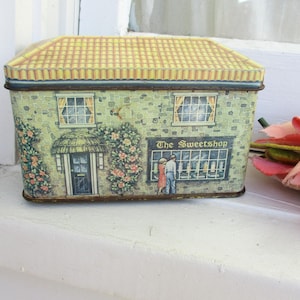 May include: A rectangular tin box designed to resemble a quaint cottage. The box features a yellow tiled roof, a green stone facade, and the words "The Sweetshop" above a shop window. Floral accents and a thatched door complete the charming design.