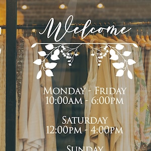May include: A white sign with black text on a glass storefront window. The sign says "Welcome" with a floral design. The sign also lists the store's hours of operation: Monday - Friday 10:00 AM - 6:00 PM, Saturday 12:00 PM - 4:00 PM, and Sunday Closed.