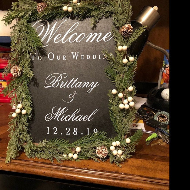 以下が含まれることがあります： 黒いチョークボードの看板に白い文字で「Brittany & Michael 12.28.19 結婚式へようこそ」と書かれています。看板は緑の松の枝と白いベリーのガーランドで飾られています。