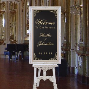 以下が含まれることがあります： 黒と金の結婚式の看板が置かれた白いイーゼル。看板には「Welcome to our wedding Heather & Johnathan 04.25.18」と書かれている