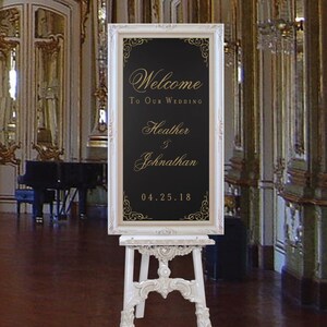 以下が含まれることがあります： 金色の文字で「私たちの結婚式へようこそ Heather & Johnathan 04.25.18」と書かれた黒い額縁の看板がついた白いイーゼルです。