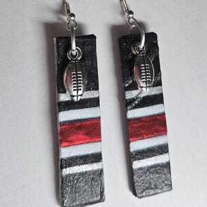 May include: A pair of rectangular drop earrings featuring a striped pattern in black, white, and red. Each earring is accented with a small, silver-toned metal charm shaped like an American football, suspended from a silver-toned hook wire.