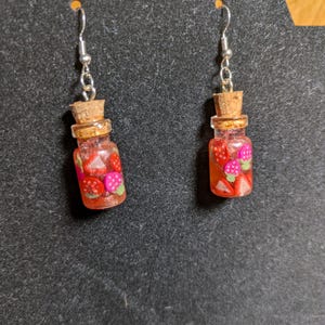 May include: A pair of dangle earrings featuring miniature glass bottles filled with red and pink strawberry charms. Each bottle has a cork stopper and is suspended from a silver-colored fish hook ear wire. The earrings are displayed against a dark gray background.